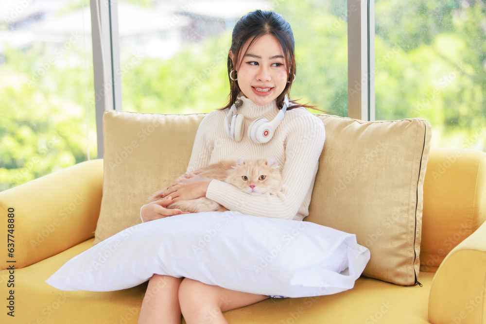 Asian young beautiful female teenager girl owner in turtleneck sweater wearing headphones listening to music sitting smiling on cozy sofa holding cute little orange long hair domestic cat on pillow Asian young beautiful female teenager girl owner in turtleneck sweater wearing headphones listening to music sitting smiling on cozy sofa holding cute little orange long hair domestic cat on pillow