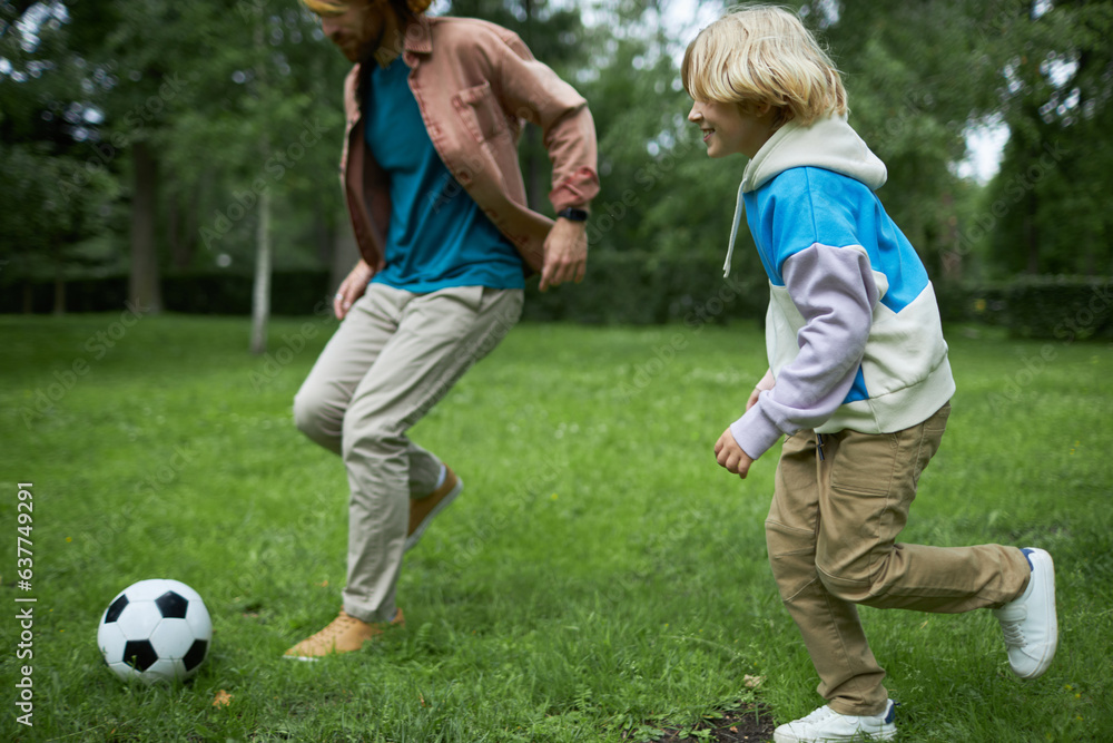 Obraz premium Candid shot of father and son running in park together and playing football on green grass