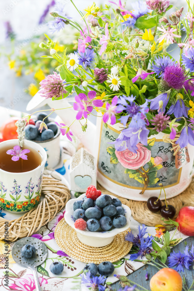Still life with meadow flowers bouquet, berries and cup of tea in natural light, vivid wild flowers, berries on table with wicker small trays, close up view