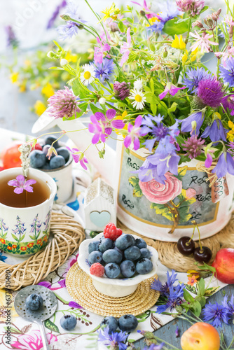 Still life with meadow flowers bouquet  berries and cup of tea in natural light  vivid wild flowers  berries on table with wicker small trays  close up view