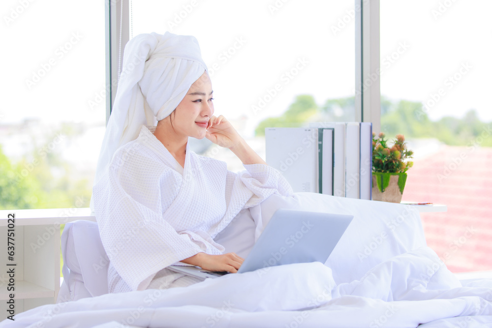 Asian young pretty sexy female teenager girl in bathrobe ready for relaxing spa on bed sitting smiling looking outside while surfing browsing internet via laptop notebook computer online in bedroom. Asian young pretty sexy female teenager girl in bathrobe ready for relaxing spa on bed sitting smiling looking outside while surfing browsing internet via laptop notebook computer online in bedroom.