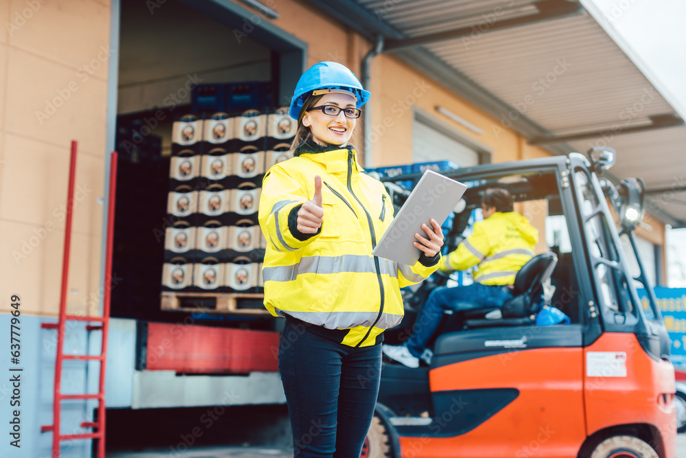 woman in industrial gear standing in front of a forklift transporting goods from a warehouse woman in industrial gear standing in front of a forklift transporting goods from a warehouse