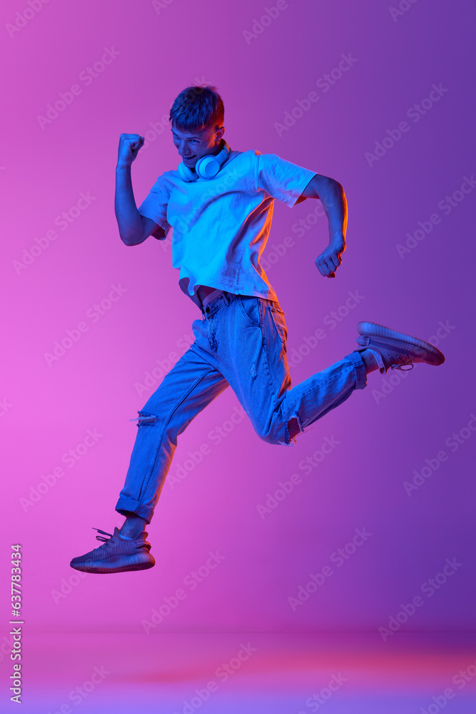 Full-length image of young guy in casual clothes and headphones jumping over gradient pink purple background in neon light. Concept of human emotions, youth, lifestyle, fashion, facial expressions, ad