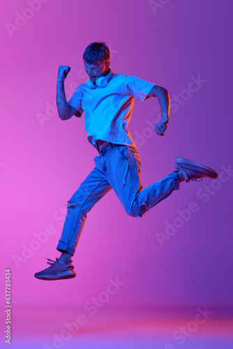 Full-length image of young guy in casual clothes and headphones jumping over gradient pink purple background in neon light. Concept of human emotions, youth, lifestyle, fashion, facial expressions, ad