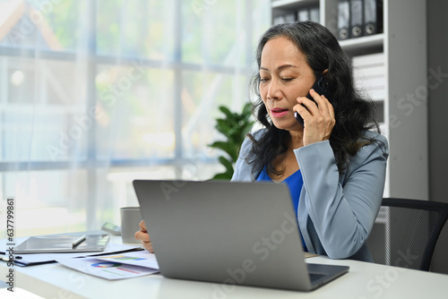 Serious middle aged entrepreneur woman talking on cellphone and using laptop at office desk