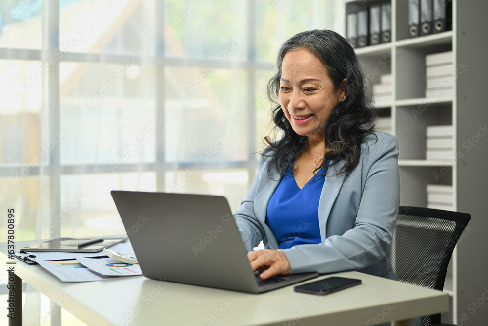 Portrait of positive mature businesswoman using laptop computer, typing, working in modern office Portrait of positive mature businesswoman using laptop computer, typing, working in modern office
