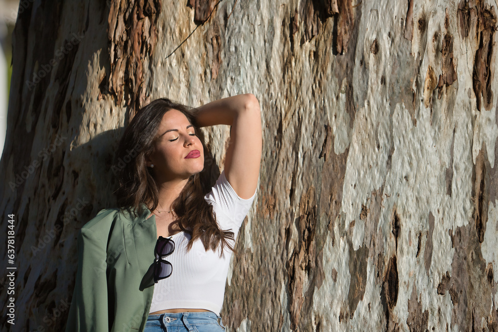 Young and beautiful woman, Hispanic, brown hair, with green jacket and jeans, eyes closed, alone, relaxed and calm, leaning against the trunk of a huge tree. Concept of solitude, relax, calm. Young and beautiful woman, Hispanic, brown hair, with green jacket and jeans, eyes closed, alone, relaxed and calm, leaning against the trunk of a huge tree. Concept of solitude, relax, calm.