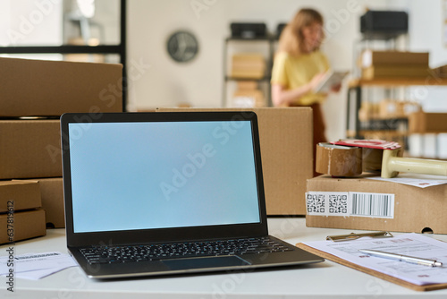 White screen of laptop standing on desk among packed cardboard boxes and financial documents on background of young female manager