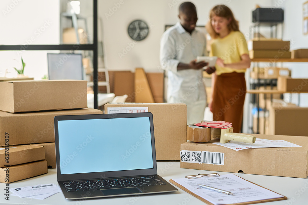 Laptop with white empty screen standing on desk among packed boxes and documents against two young intercultural managers using tablet