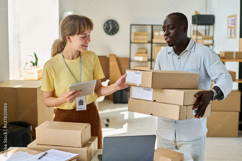 Happy young woman with tablet checking personal data of clients on cardboard boxes held by her African American male colleague Happy young woman with tablet checking personal data of clients on cardboard boxes held by her African American male colleague