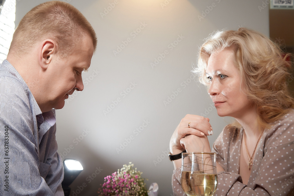 A couple at a table, where the man is sitting and reading on his cell phone while the woman looks sadly at him. Divorce, quarrel, infidelity, loneliness, discord, dissatisfaction - obrazy, fototapety, plakaty 