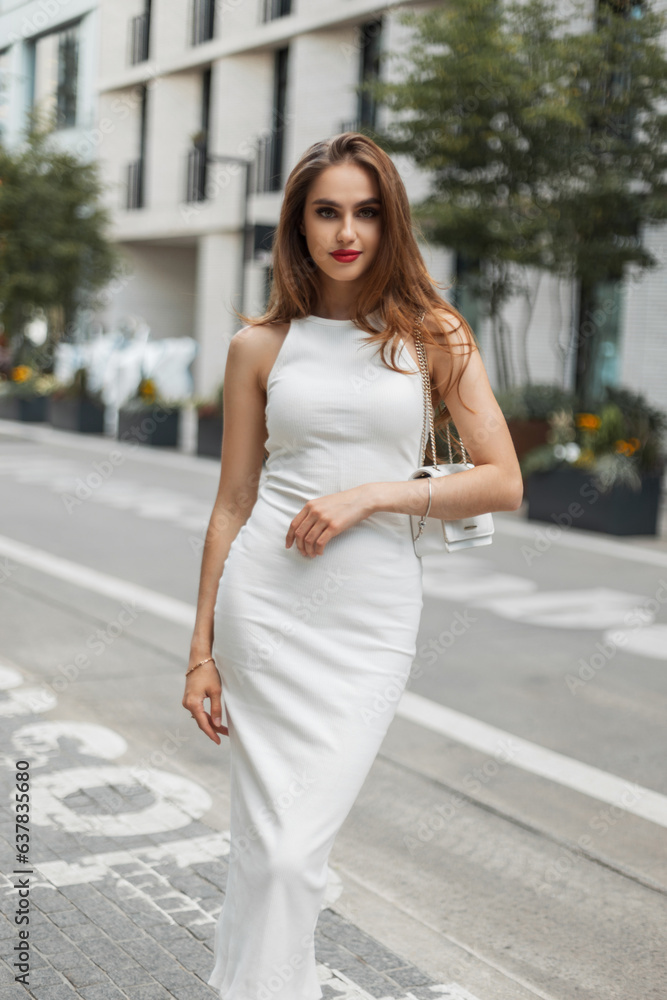 Fashionable chic beauty woman model with red lips in white elegant fashion dress with bag walks on the street. Pretty lady Fashionable chic beauty woman model with red lips in white elegant fashion dress with bag walks on the street. Pretty lady