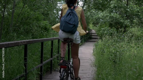 Young brown hair woman with backpack riding bicycle on park, back view. Camera moving forward after her, slow motion effect