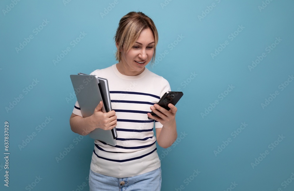 confident smiling blonde girl uses app on smartphone holding laptop in hands on blue background with copy space confident smiling blonde girl uses app on smartphone holding laptop in hands on blue background with copy space