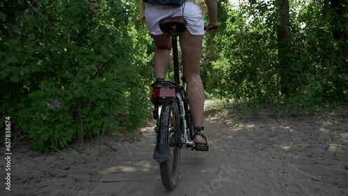 Close-up of a woman's legs in white shorts on a bicycle alone in a summer park near the sea. Sunny day, sand and grass. Unrecognizable girl on a bike, healthy lifestyle, aesthetic footage.