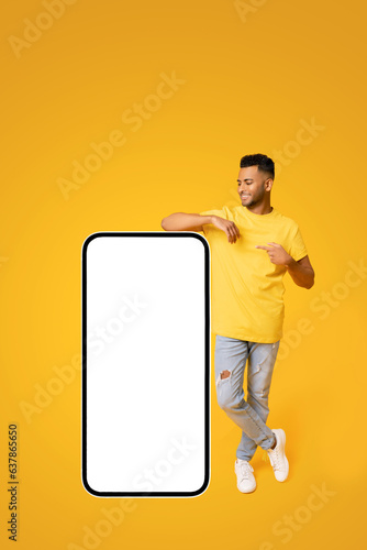 Best deal promotion. Smiling indian teenage guy leaning on huge smartphone with blank screen, pointing finger at empty display and smiling, arab man advertising new app or website, mockup