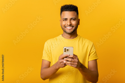 Portrait of cheerful young Indian guy sending message on mobile phone over yellow background. Happy Arab man using smartphone, enjoying chatting online, messaging in social networks © Vadim Pastuh