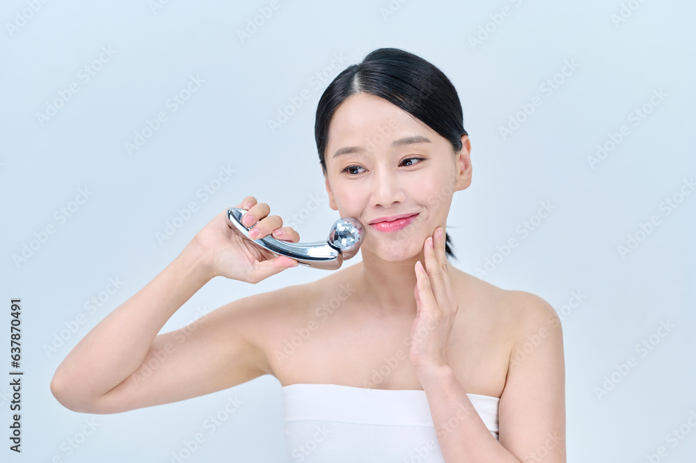 아시아 한국의 검은머리의 젊은 여자 모델이 끈 나시를 입고 은색 마사지 롤러를 얼굴 목 어깨에 대고 포즈를 취하고 있다. 아시아 한국의 검은머리의 젊은 여자 모델이 끈 나시를 입고 은색 마사지 롤러를 얼굴 목 어깨에 대고 포즈를 취하고 있다.