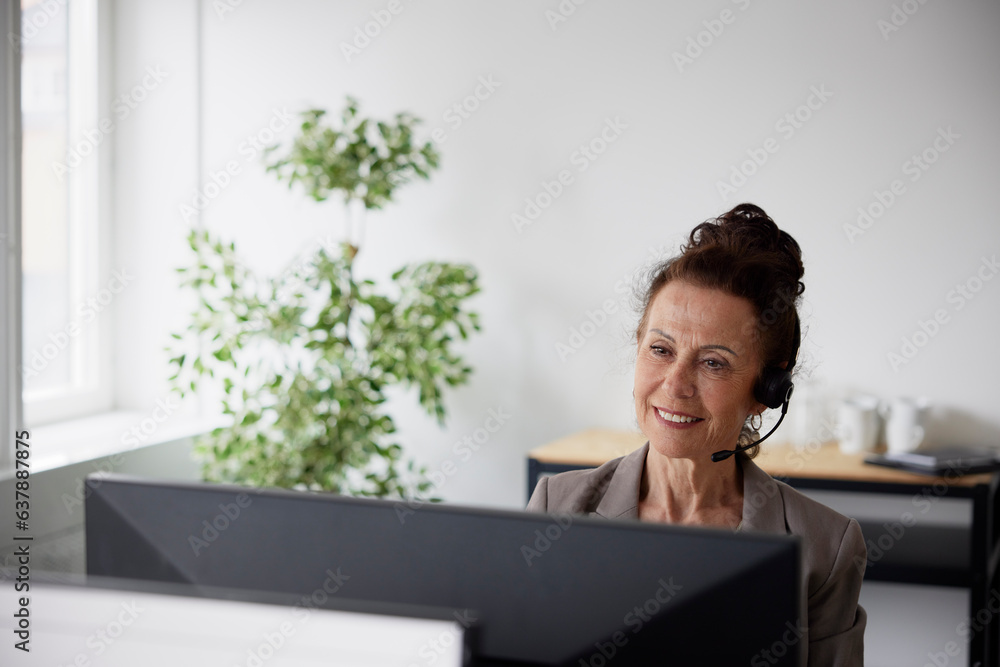 Smiling mature businesswoman using headset in office in front of computer screen Smiling mature businesswoman using headset in office in front of computer screen