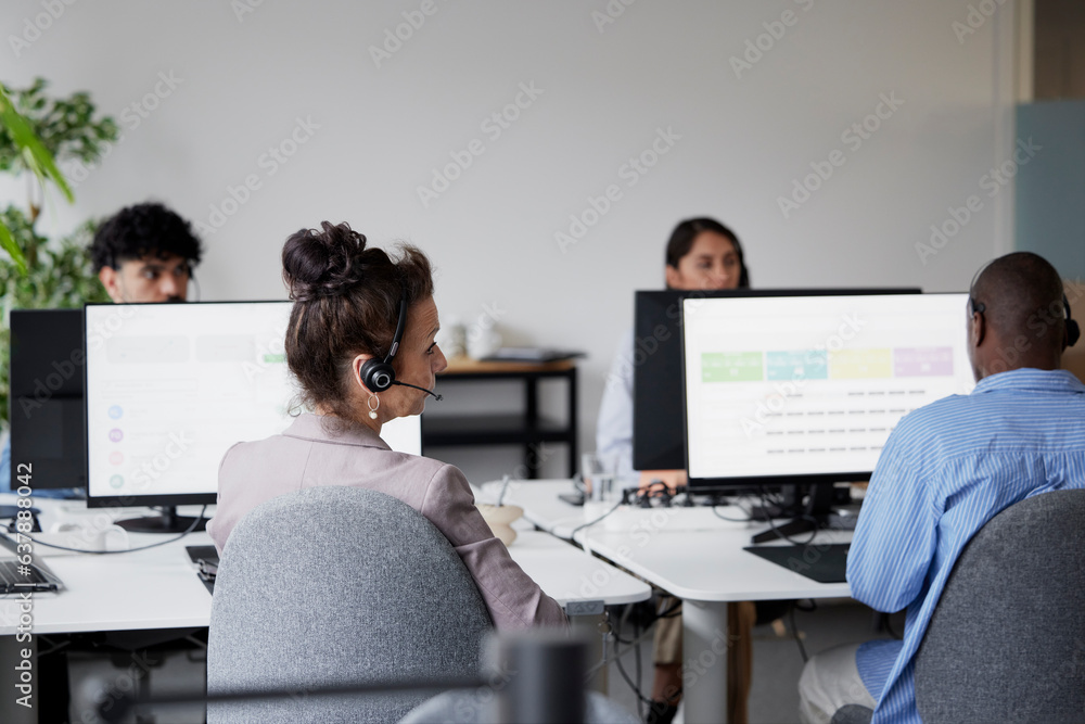 Rear view of businesswoman using headset in office in front of computer screen Rear view of businesswoman using headset in office in front of computer screen