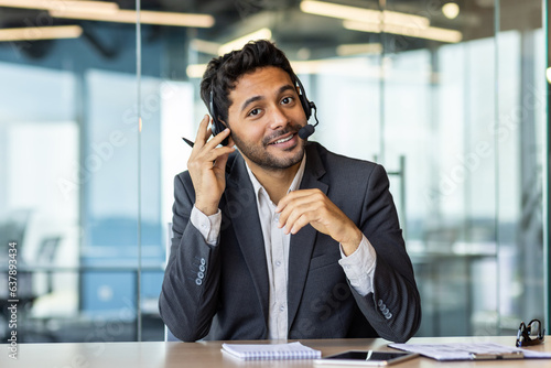 Portrait of young successful arab businessman, man with headset phone for video call smiling and looking at camera, employee senior customer service manager at workplace inside office.