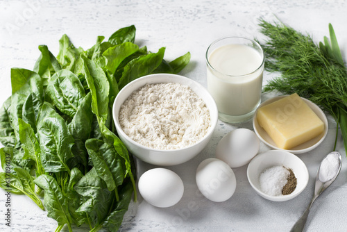 Ingredients: spinach, sorrel, flour, eggs, yogurt, cheese, spices, baking powder and greens for cooking a delicious vegetarian pie or cupcake on a light blue background, top view