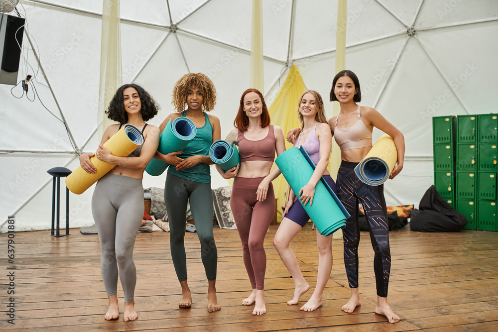 Fototapeta premium full length of happy multiethnic women in sportswear standing with yoga mats and looking at camera