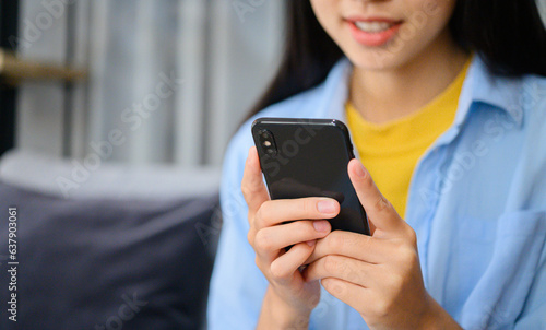 Smiling young asian woman using mobile phone checking social media,app playing game, shopping online, ordering delivery relax while sitting on a couch at home with laptop computer