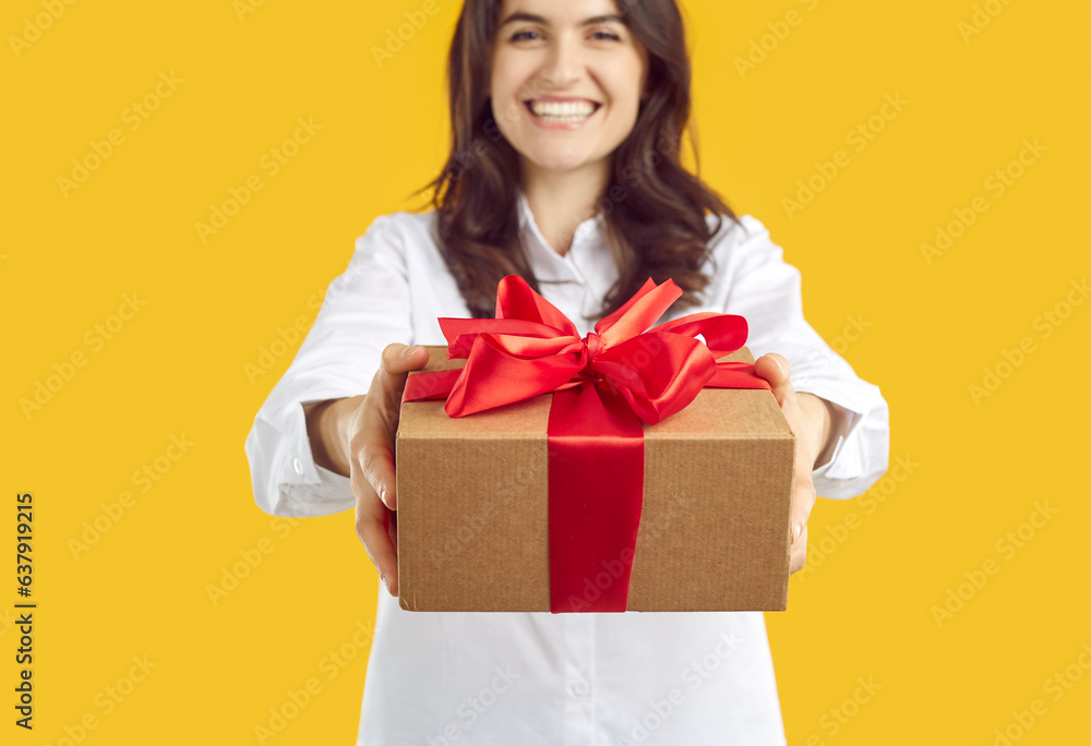 Cheerful joyful young woman holds out gift box tied with red ribbon with bow to camera. Close up of gift box in hands of woman on yellow background. Concept of holiday gifts. Selective focus. Cheerful joyful young woman holds out gift box tied with red ribbon with bow to camera. Close up of gift box in hands of woman on yellow background. Concept of holiday gifts. Selective focus.