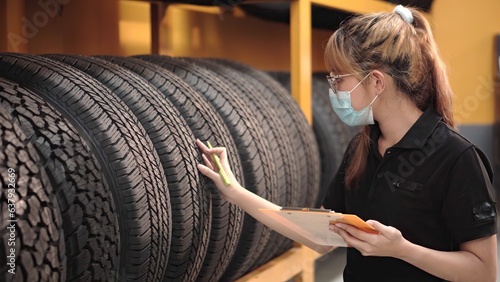 Worker young asia woman wearing face medical mask is checking quality of car tires and checking the stock of car tires in auto repair shop store during covid 19 epidemic.