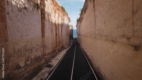 Aerial drone flying over asphalt road and high rock mountain walls on both sides. Bali Pandawa beach approaching between sandstone cliff, water and blue sky. Nature and travel concept