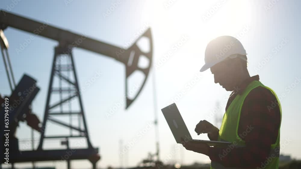 oil business. a worker works next to an oil pump holding a laptop. industry business lifestyle oil and gas concept. engineer studying the level of oil production on a laptop silhouette at sunset oil business. a worker works next to an oil pump holding a laptop. industry business lifestyle oil and gas concept. engineer studying the level of oil production on a laptop silhouette at sunset