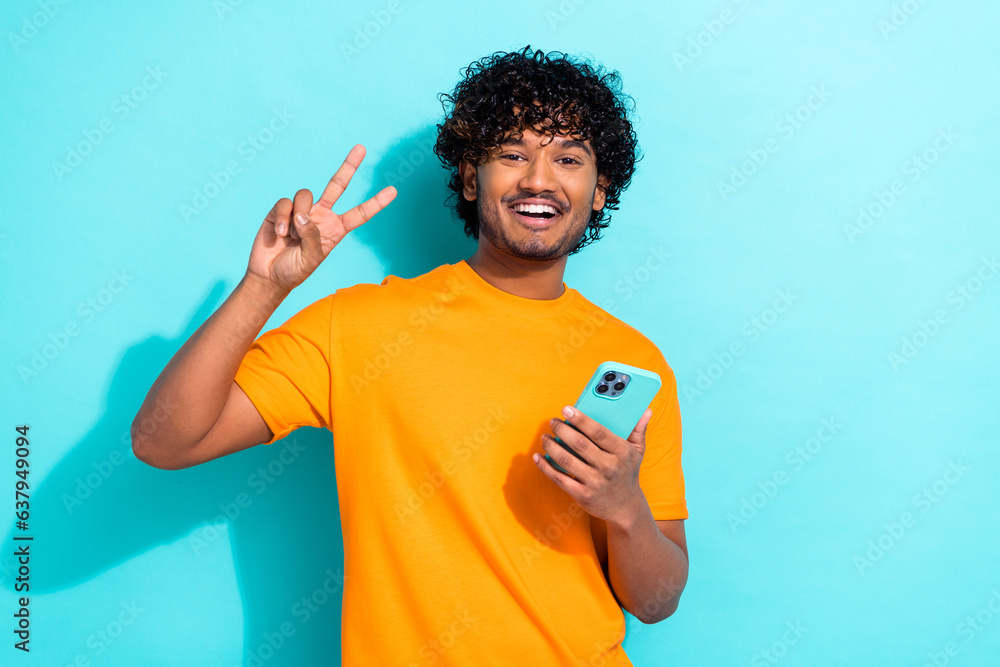 Photo of funky positive handsome man toothy smile hold smart phone hand fingers demonstrate v-sign isolated on teal color background Photo of funky positive handsome man toothy smile hold smart phone hand fingers demonstrate v-sign isolated on teal color background
