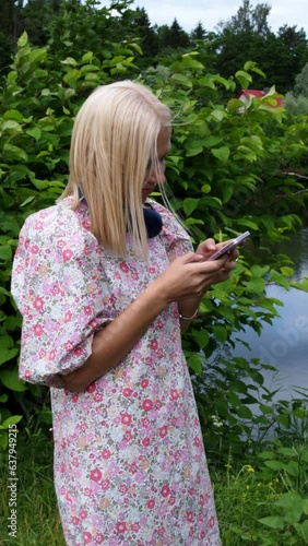 Young woman in a floral dress stands by the lake and looks at her smartphone