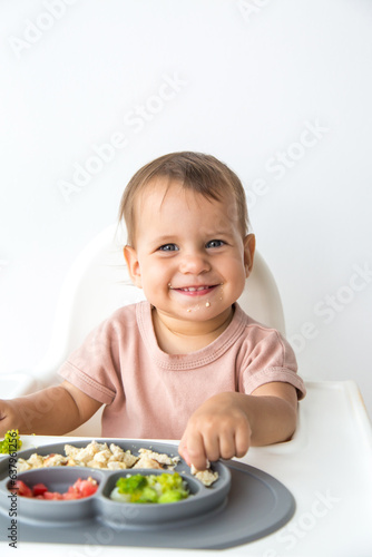 vertical photo of a cute beautiful baby 1 year old sitting in a highchair and eating complementary foods, smiling and looking at the camera on a white background
