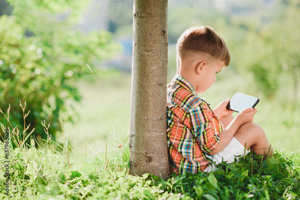 A beautiful child sits on the grass and looks at the phone in the summer at sunset. Cute boy with mobile in hand A beautiful child sits on the grass and looks at the phone in the summer at sunset. Cute boy with mobile in hand