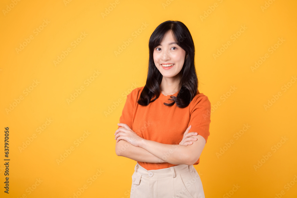 Embrace confidence with a young Asian woman in her 30s, donning an orange shirt, displaying crossed arm sign gesture on yellow background. Embrace confidence with a young Asian woman in her 30s, donning an orange shirt, displaying crossed arm sign gesture on yellow background.