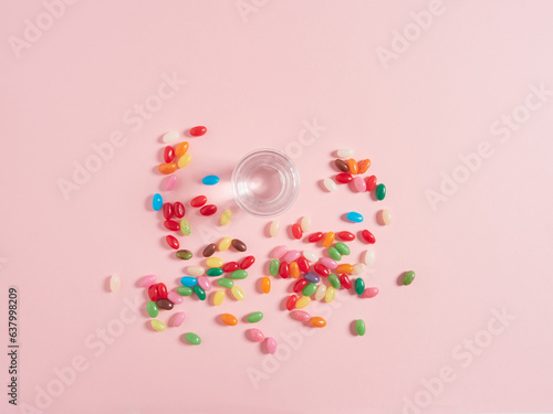 View from above glass of water and jelly beans as colored pills scattered on pink background. Concept of vitamins and food supplement.