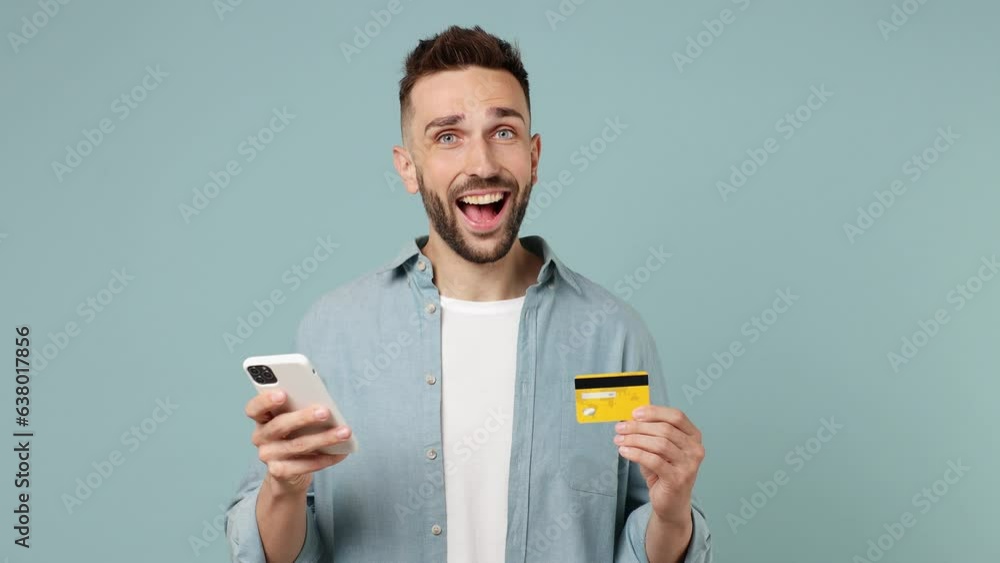 Young fun man wear shirt white t-shirt use mobile cell phone app hold in hand credit bank card doing online shopping order delivery to home isolated on plain pastel light blue cyan background studio Young fun man wear shirt white t-shirt use mobile cell phone app hold in hand credit bank card doing online shopping order delivery to home isolated on plain pastel light blue cyan background studio