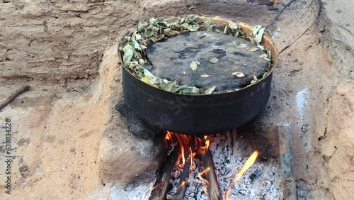 A large black pot sits over a roaring open fire, surrounded by an earthy pit. Flames lick the bottom of the pot, which is covered with green leaves, typical of rural outdoor cooking techniques in Indi photo