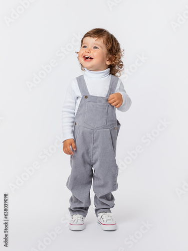 A cheerful laughing Caucasian toddler of 2 years old with curly hair in a gray jumpsuit and a white turtleneck stands tall and looks up. Expressive look