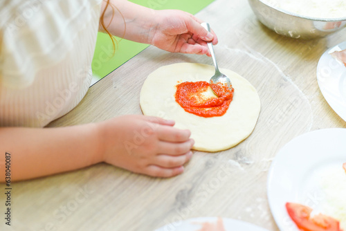 Children s hands with a spoon spreading ketchup on the dough. Child little cook prepares pizza