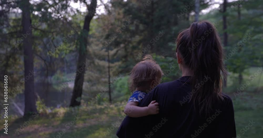Mother walking with toddler girl in arms through forest on sunny morning - from behind Mother walking with toddler girl in arms through forest on sunny morning - from behind