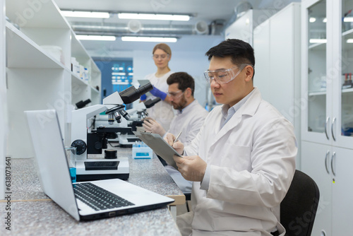 A team of scientists and doctors work in the laboratory. Asian man writing results and sitting at laptop  male and female colleagues examining under microscope.