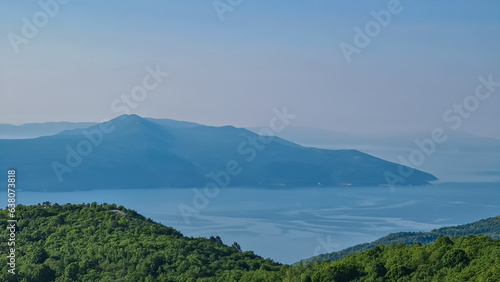 A panoramic view on the Mediterranean Sea in Croatia. The sun has just risen, leaving the sky still a bit orange. The hills are overgrown with lush trees and bushes. Early morning hiking by the sea.