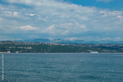 A panoramic view of the Croatian shore. There are some towns located on the shore of the Mediterranean Sea. Calm surface of the sea. Stony beach. Green hills in the back. Sunny day with puffy clouds