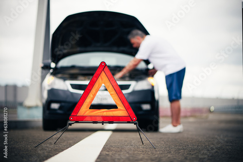 Red emergency stop sign on road. Blurred man fixing broken down car. Warning triangle on roadside. Safe traffic, emergency situation in trip. Help and service on the road.