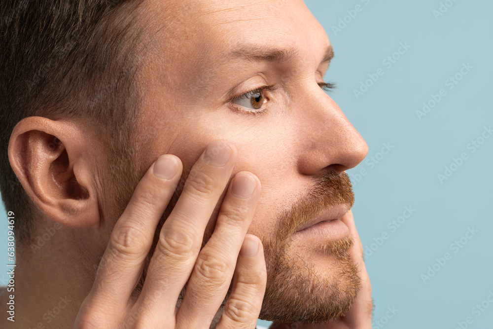 Interested man worried looks at skin on face touch cheeks think about wrinkles, anti aging cosmetic cream products, cosmetology regeneration lifting procedure against studio background. Facial surgery Interested man worried looks at skin on face touch cheeks think about wrinkles, anti aging cosmetic cream products, cosmetology regeneration lifting procedure against studio background. Facial surgery