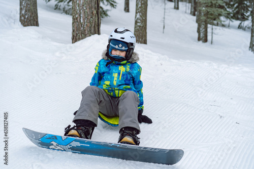 little boy sitting on snow putting his feet in snowboard bindings adjusting straps. 
