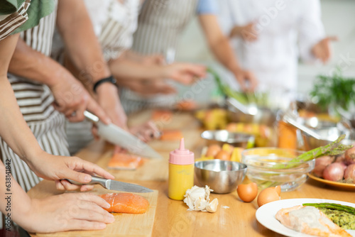 People in aprons standing at table with groceries and utensils during group cooking lesson, absorbed in cooking process, cutting fresh salmon following chef instructions. Cropped shot photo